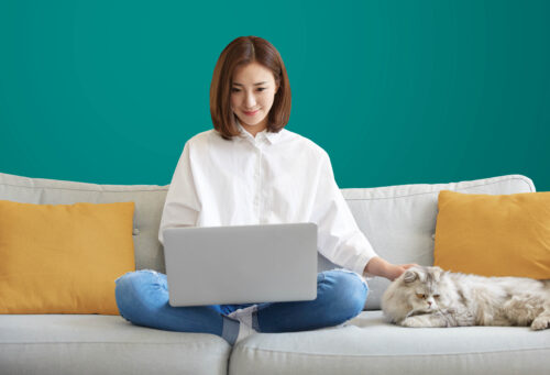 A woman sits comfortably on her couch, typing on her laptop with her cat curled up beside her