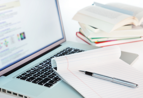 An open notebook sits atop a laptop and several books are stacked in the background