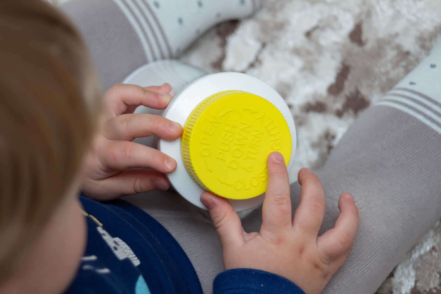 A child independently tries to open a plastic bottle of medicine with a yellow push and turn lid, close-up.