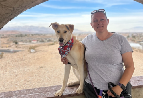 Leanne James and her dog Willow, a tan dog with one blue eye and one black eye, stand in front of a desert landscape