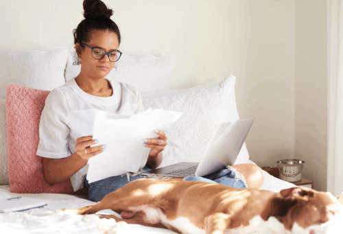 woman looking through papers with a laptop on her lap and a dog laying next to her