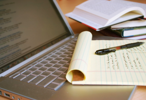 An open laptop sits on a desk with a notepad on top of the keyboard