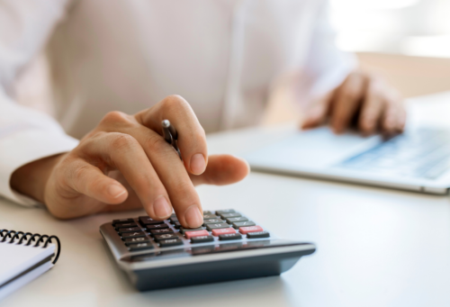 A close up of a person's hand using a calculator while sitting at a desk in front of a laptop
