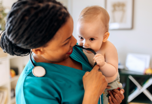 A woman wearing scrubs holds a baby who is chewing on her stethoscope