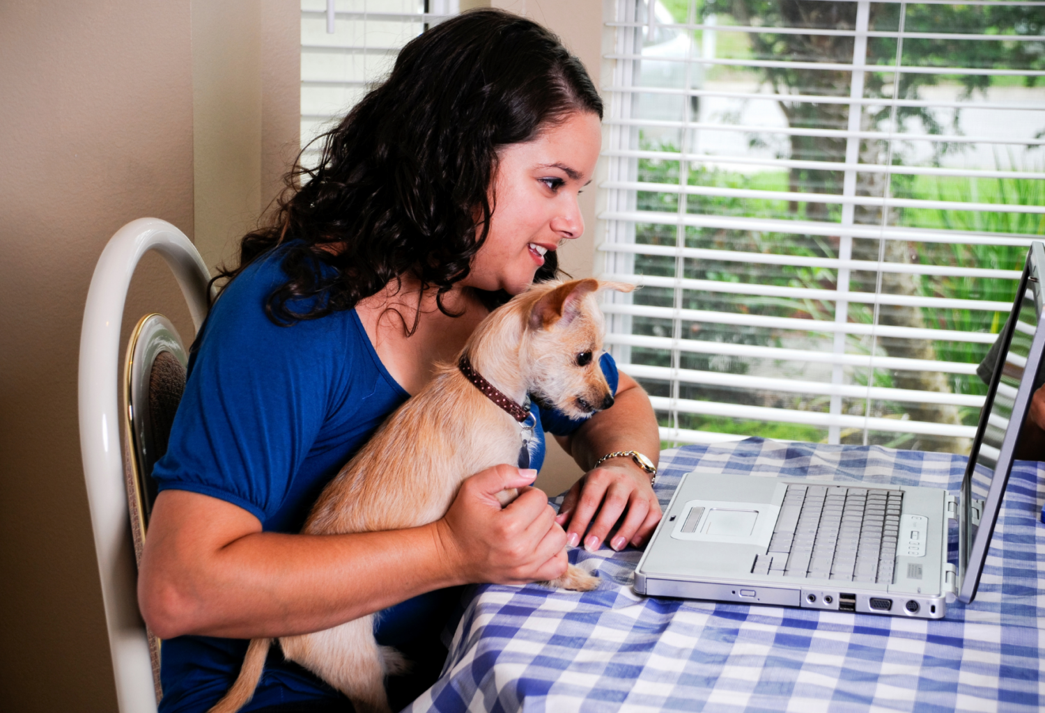 A woman sits at a table looking at her laptop with her dog